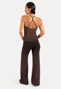 Femme aux cheveux courts et foncés portant un débardeur marron à fines bretelles et un pantalon à jambes larges, debout les mains sur les hanches, tournée vers l'arrière.