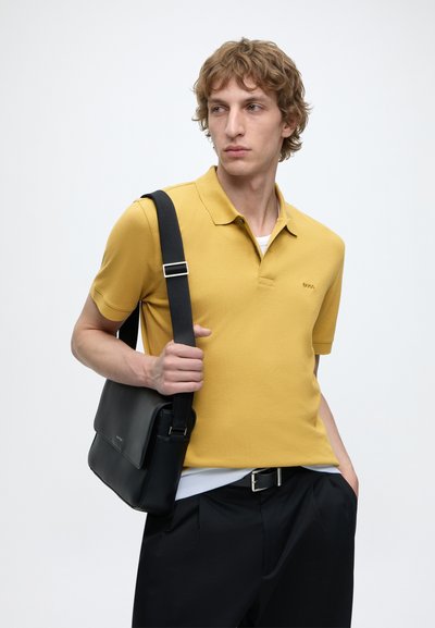 Jeune homme portant un polo jaune et un pantalon noir, portant un sac à bandoulière noir, regardant sur le côté sur un fond blanc uni.