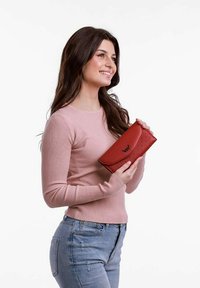 Červená ko�žená peněženka s texturovaným povrchem a zakřiveným klopou, držena v ruce. Viditelné jsou také proužkované dlouhé rukávy a světlé džíny.