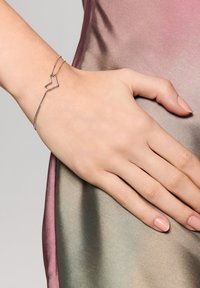 Srebrna bransoletka o kształcie serca na delikatnym łańcuszku, noszona na ręce na tle gładkiego, gradientowego jedwabnego materiału.