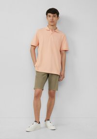 Lichtroze polo shirt met een kraag en korte mouwen, gecombineerd met kaki shorts. Model draagt witte sneakers en staat tegen een effen achtergrond.