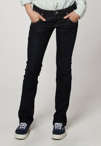 Jeans de mezclilla azul oscuro con corte slim, que cuentan con cinco bolsillos, costuras visibles en un color contrastante y un cierre de botón.