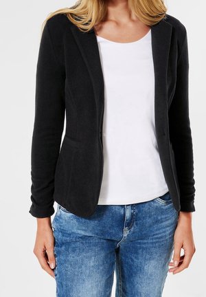 Vrouw draagt een zwarte blazer over een wit topje en blauwe spijkerbroek, staand tegen een effen lichte achtergrond.