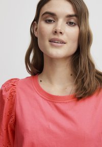 Tricou scurt cu mânecă scurtă de culoare corai, cu guler rotund; are mâneci puff dintr-un material brodat. Textură netedă, croi lejer.