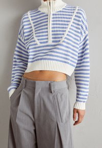 Maglione corto a maglia con righe blu e bianche, collo con mezzo zip, indossato con pantaloni grigi a gamba larga a vita alta e una sottile catena in vita.