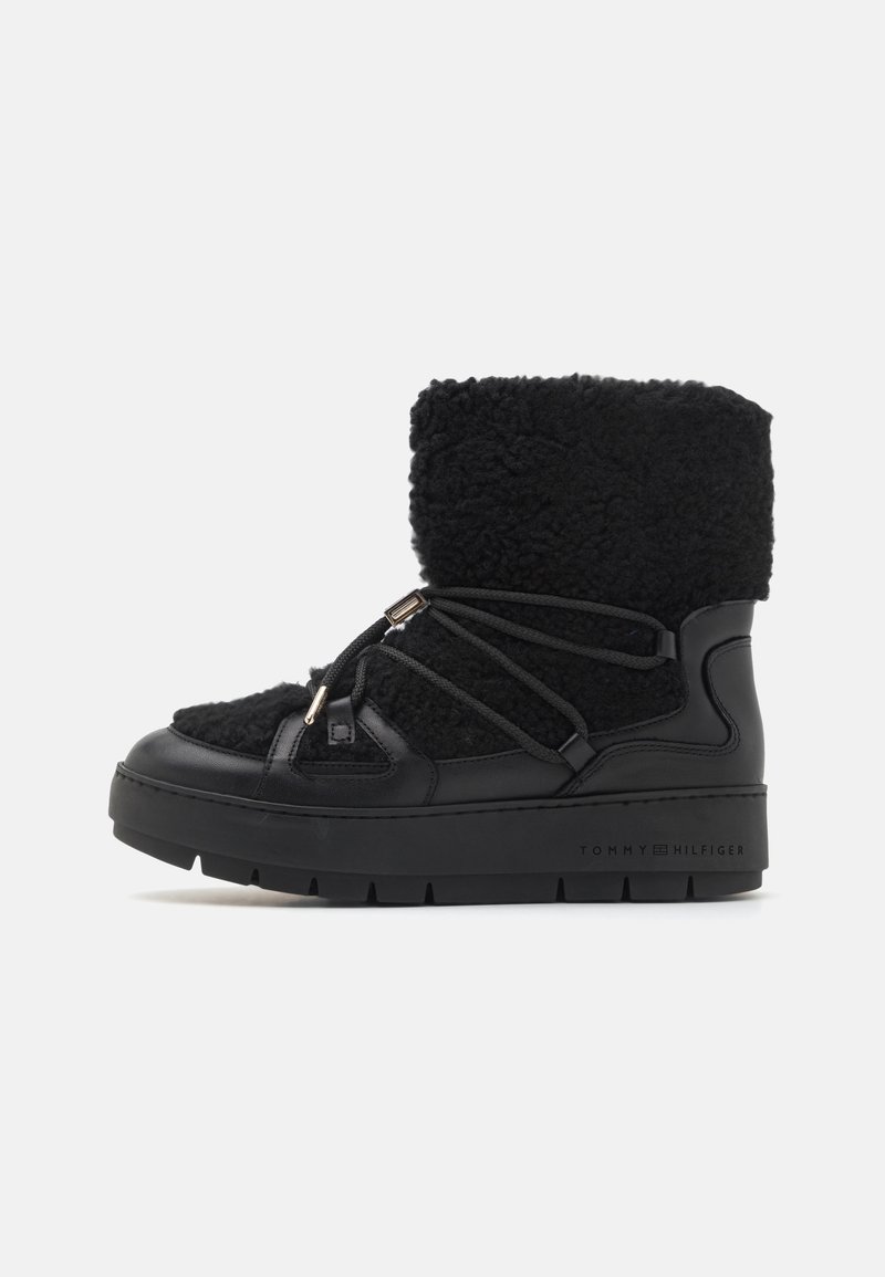 Botte d'hiver noire mi-mollet avec tige en polaire, semelle épaisse et lacets enveloppés avec accents métalliques sur fond blanc.