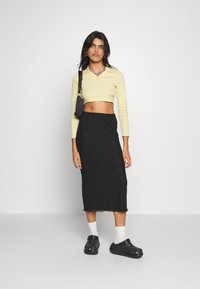 T-shirt amarelo cropped de mangas longas, saia midi preta e tamancos pretos. A t-shirt tem um decote em V e uma marcação discreta; a saia tem uma textura suave.