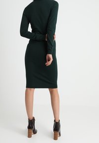 Robe tricotée verte avec des manches longues, de forme ajustée et texture côtelée. Portée avec des bottines noires à talons empilés.