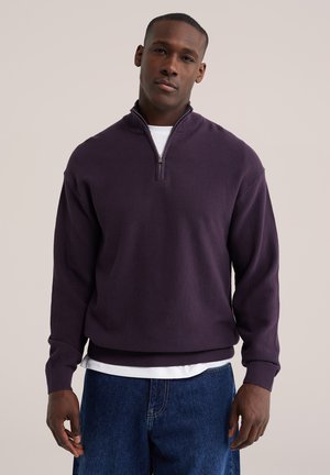 Hombre con un suéter de cremallera corta de color púrpura oscuro sobre una camisa blanca y vaqueros azules, de pie contra un fondo claro y liso.
