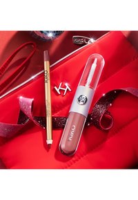 Kosmetikkartiklene inkluderer en gull leppekonturpen og en klar leppeglans i en hvit beholder, plassert på en rød satengpose med glitrende bånd.