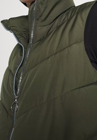 Pikowana kamizelka w oliwkowej zieleni z wysokim kołnierzem, zapinana na zamek błyskawiczny z ozdobnymi srebrnymi akcentami na zamku. Gładka, fakturowana tkanina.