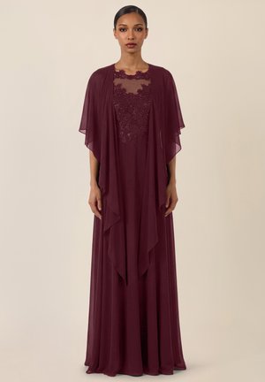 Frau in einem bodenlangen burgunderfarbenen Kleid mit Spitzenmieder und durchsichtiger capeartiger Überlagerung, stehend vor einem schlichten beigen Hintergrund.