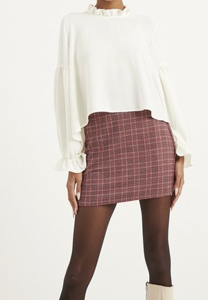 Kobieta w białej, falbaniastej bluzce z długimi bufkami na rękawach, burgundowej kratkowanej mini spódnicy, czarnych rajstopach i beżowych botkach za kostkę.