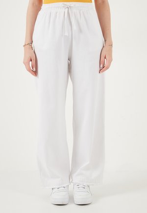 Persoană care poartă pantaloni albi largi cu șnur și adidași albi, stând cu mâinile relaxate pe lângă corp.