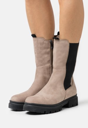 Bottines en daim beige clair avec des panneaux élastiques noirs sur les côtés et une semelle extérieure en caoutchouc noir texturé. Comprend une fermeture éclair sur le côté pour un enfilage facile.