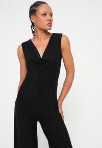 Mono negro sin mangas con escote drapeado, cuerpo ajustado y piernas anchas, hecho de una tela texturizada y brillante.
