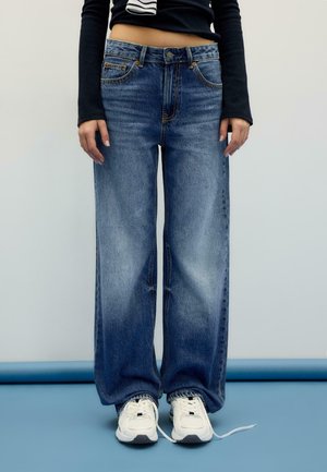 Person iført blå vidde jeans med hvide sneakers og en sort cropped langærmet top, stående på en blå gulv mod en ensfarvet baggrund.
