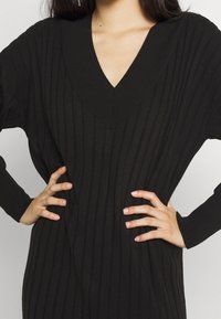 Svart ribbad stickad klänning med djup V-ringning och långa ärmar. Materialet har en vertikal textur och en avslappnad passform.