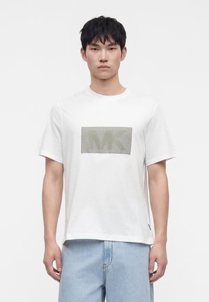 Νέος άνδρας με λευκό T-shirt κοντών μανικιών με γκρίζο λογότυπο MK στο στήθος και ανοιχτό μπλε τζιν, στέκεται μπροστά σε απλό φόντο.