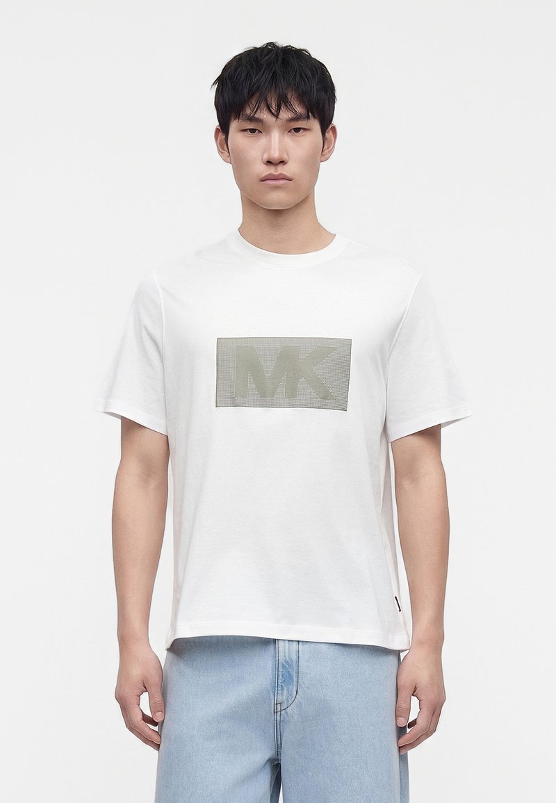 Νέος άνδρας με λευκό T-shirt κοντών μανικιών με γκρίζο λογότυπο MK στο στήθος και ανοιχτό μπλε τζιν, στέκεται μπροστά σε απλό φόντο.