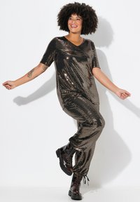 Bronzen jumpsuit met pailletten, korte mouwen, V-hals, losse pasvorm en taps toelopende pijpen, gecombineerd met zwarte veterschoenen.