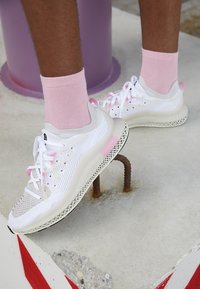 White athletic shoes with pink accents, breathable mesh upper, textured midsole, and a patterned outsole. Pink socks visible above the shoe.