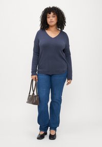 Mornarsko V-neck pulover iz mehkega pletenega materiala, nosi se z modrimi kavbojkami. Temna torba z dvema ročajema, črne čevlje. Krepki lasje. Minimalistična ozadje.