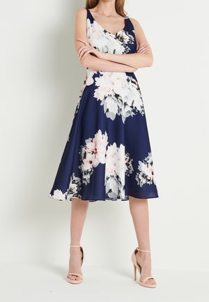 Femme portant une robe midi bleu marine à imprimé floral blanc et rose, sans manches, et des sandales à talons hauts couleur nude, debout les bras croisés.