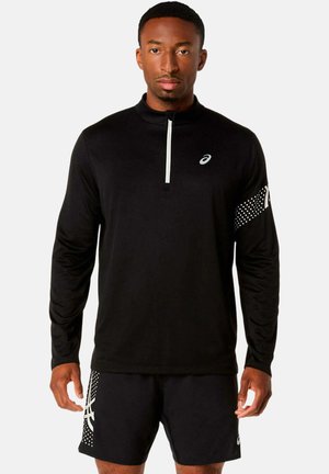 Hombre vistiendo una camiseta deportiva negra de manga larga con cremallera blanca y pantalones cortos negros con logo blanco y patrón de puntos, de pie frente a un fondo liso.