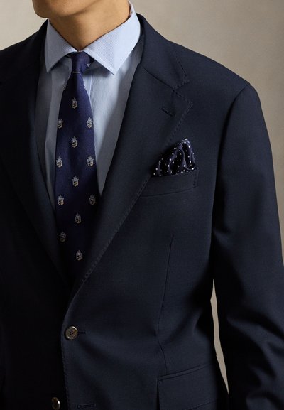 Μπλε μπουφάν ναυτικού με ένα κουμπί και πέτο με εγκοπές, φορεμένο πάνω από ένα ανοιχτό γαλάζιο ριγέ πουκάμισο, με πουά μπλε γραβάτα και μαντηλάκι τσέπης με πουά μοτίβο.