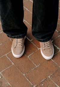 Baskets en daim beige avec semelles blanches portées avec un pantalon noir, debout sur un pavé en briques marron.