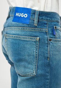 Μπλε τζιν παντελόνι με μπλε δερμάτινη ετικέτα με το "HUGO" σε λευκό, ορατή ραφή και κλασικό σχέδιο πέντε τσέπες.