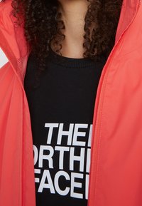 Rode windbreaker jas met een mesh voering, gecombineerd met een zwarte trui met opvallende witte tekst. Close-up view, focus op de stoftextuur en kleur.