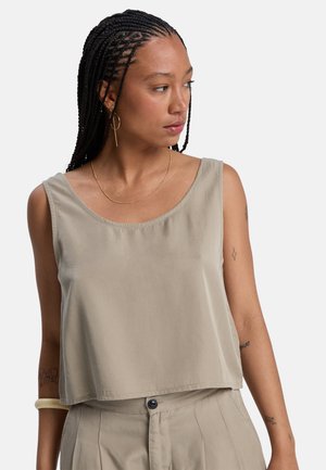 Vrouw met gevlochten haar, gekleed in een beige mouwloos topje en bijpassende high-waisted broek, kijkt naar links, met minimale sieraden en zichtbare tatoeages.