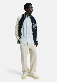 Giacca varsity in lana blu navy con maniche bianche, camicia a righe azzurro chiaro con bottoni, pantaloni beige e sneakers crema. Presenta una grande lettera "G."