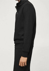 Felpa nera con zip in cotone misto, caratterizzata da un collo alto e polsini a costine. Tessuto liscio e design aderente.