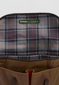 Το εσωτερικό μιας καφέ καμβά τσάντας διαθέτει μοτίβο σε γκρι, μαύρο και πορτοκαλί καρό. Ένδειξη πράσινου χρώματος αναγράφει "Η Barbour αγαπάει και τον Paul Smith."