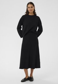 Žena stoji s rukama iza leđa, noseći crnu dugoslojnu majicu, odgovarajuću midi suknju i crne mokasine, na jednoličnoj pozadini.