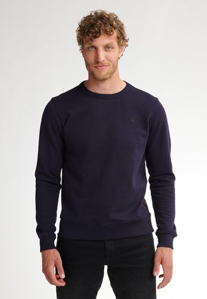 Man met krullend haar, gekleed in een marineblauwe crewneck trui en zwarte broek, staand tegen een effen witte achtergrond.