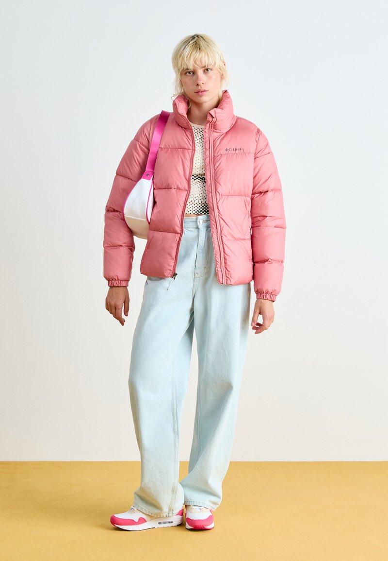 Veste matelassée rose avec un col montant et des poignets élastiques, portée sur un top noir à pois, associée avec un jean bleu clair à jambes larges et des baskets avec des accents roses.