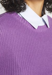 Fioletowy sweter dziergany o fakturowanej powierzchni, z okrągłym dekoltem i prążkowymi krawędziami, noszony na białej koszuli z kołnierzykiem.