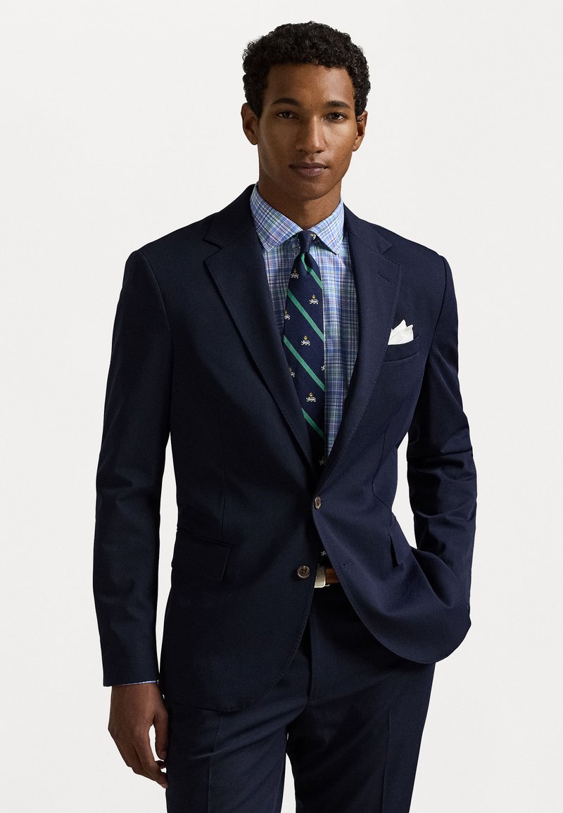 Blazer azul marinho com dois bolsos frontais, combinado com uma camisa azul xadrez e uma gravata com riscas verdes, apresentando pequenos padrões. Lenço de bolso branco incluído.