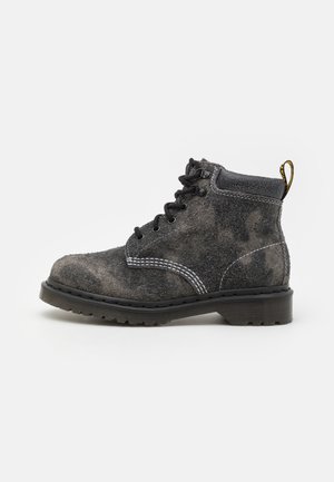 Bottine en daim gris à lacets avec semelle en caoutchouc noire, coutures blanches et languette jaune à l’arrière.