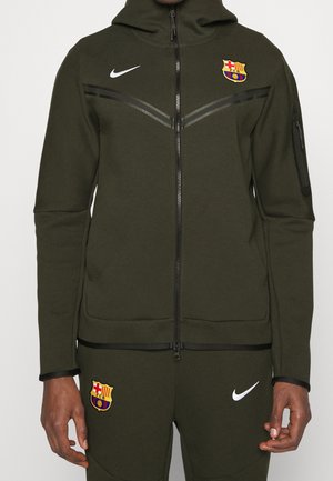 Ciemnozielony dres Nike z logo FC Barcelona na piersi i udzie, kurtka na zamek z przodu oraz kieszeń na zamek na lewym rękawie.