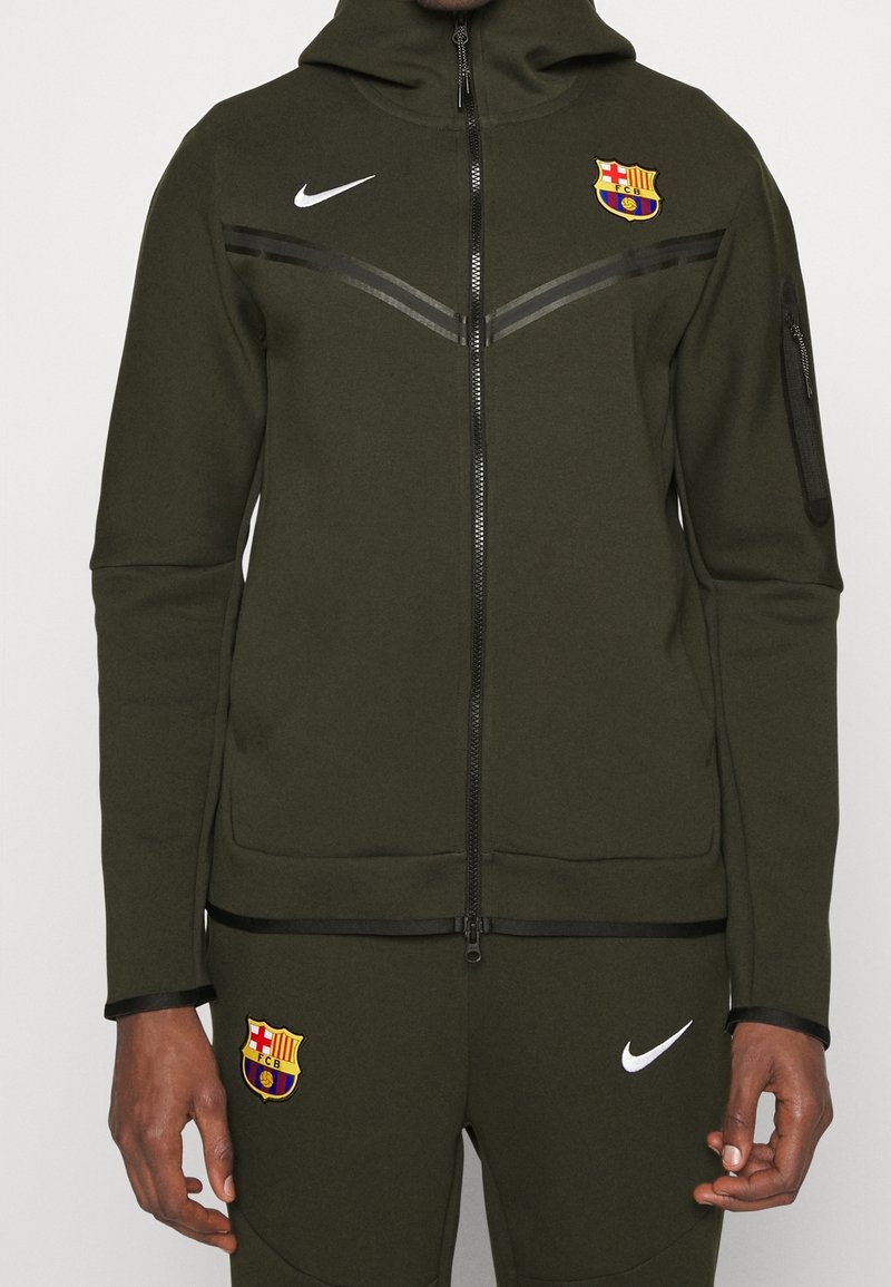 Survêtement Nike vert foncé avec logos du FC Barcelone sur la poitrine et la cuisse, veste zippée à l'avant et poche zippée sur la manche gauche.