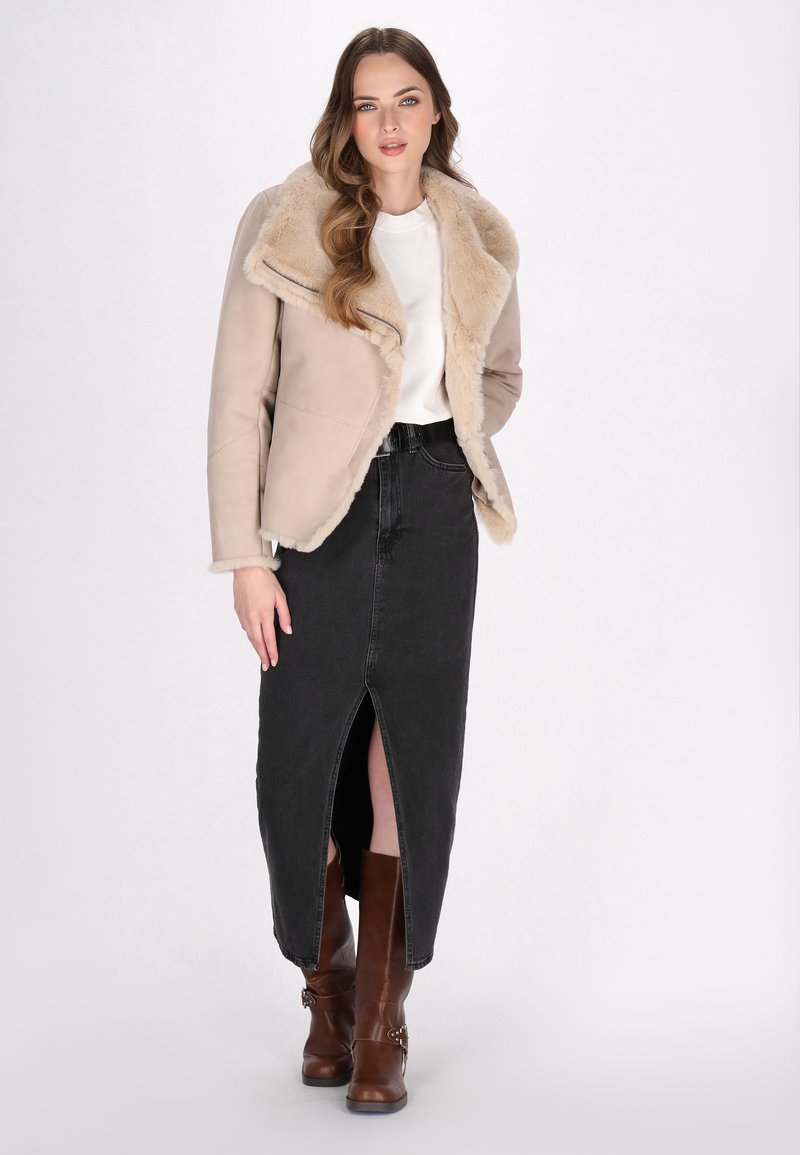 Veste en fausse fourrure beige avec grands revers, associée à une jupe en denim noire à fente avant et des bottes à talons marron.