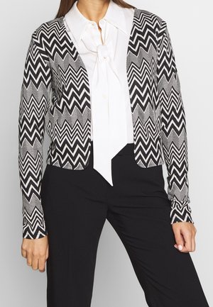 Cardigan a zigzag in bianco e nero, con maniche lunghe, apertura frontale, e un tessuto leggero e testurizzato. Abbinato a una camicetta bianca.