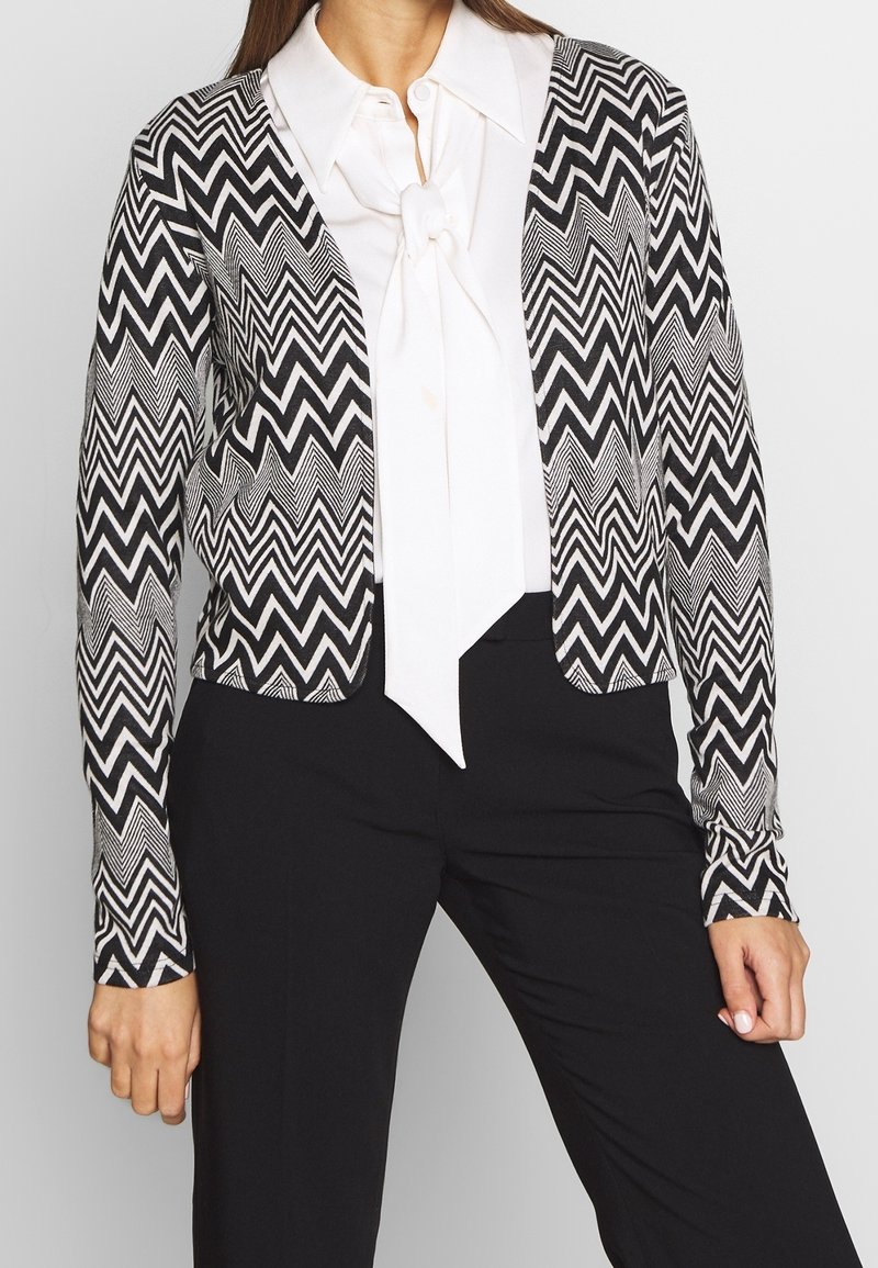 Svart och vit zigzag-mönstrad cardigan med långa ärmar, öppen fram och ett lätt, texturerat tyg. Matchas med en vit blus.
