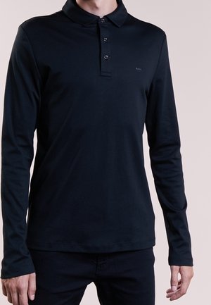 Personne portant un polo noir ajusté à manches longues avec trois boutons, debout devant un fond clair uni.