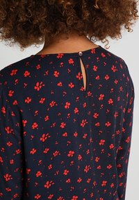 Personne aux cheveux bouclés portant une robe bleu marine à motif floral rouge, montrant l'encolure dos avec une petite fermeture à bouton et une ouverture en trou de serrure.